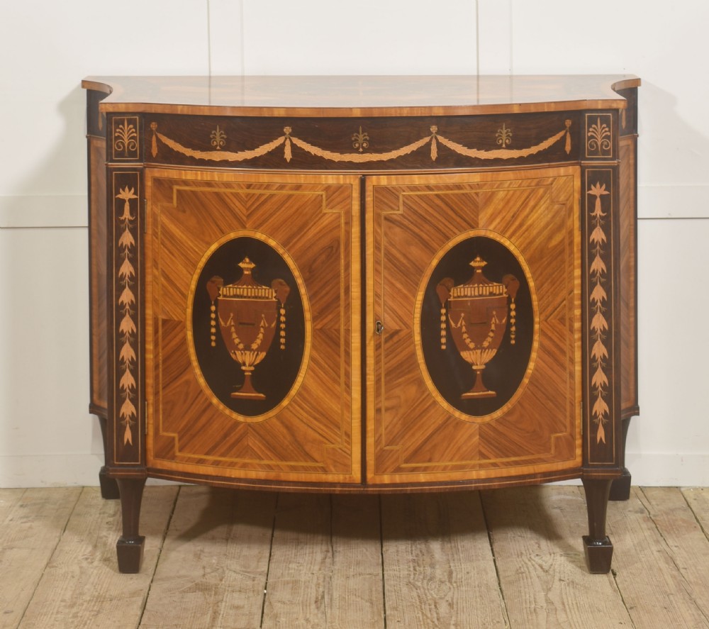 A George III style satinwood and rosewood marquetry sideboard featuring oval cabinet doors with inlaid urn medallions, floral swags, and tapered square legs.