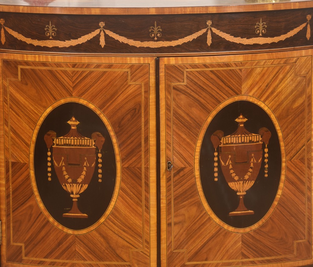 A George III style satinwood and rosewood marquetry sideboard featuring oval cabinet doors with inlaid urn medallions, floral swags, and tapered square legs. View IV