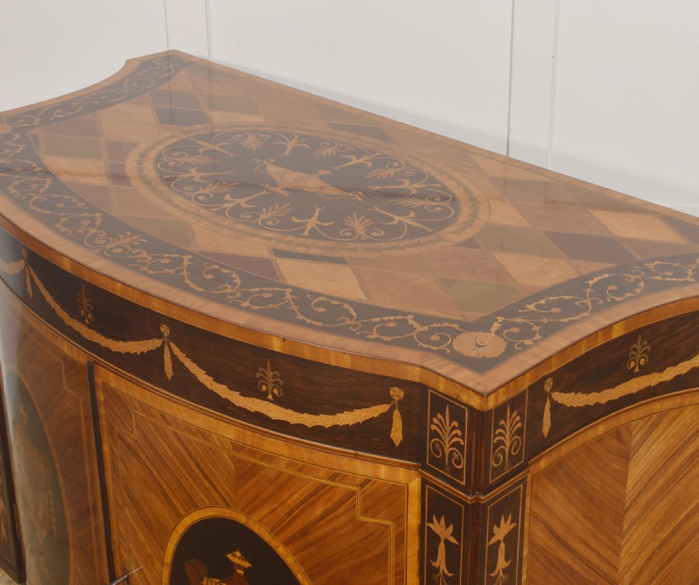 A George III style satinwood and rosewood marquetry sideboard featuring oval cabinet doors with inlaid urn medallions, floral swags, and tapered square legs. View X