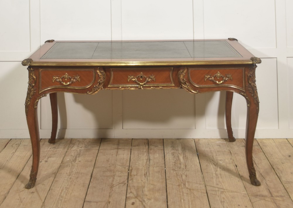 A French Louis XV style wooden bureau plat featuring three frieze drawers, gilt-bronze ormolu mounts, cabriole legs, and a green leather writing surface.