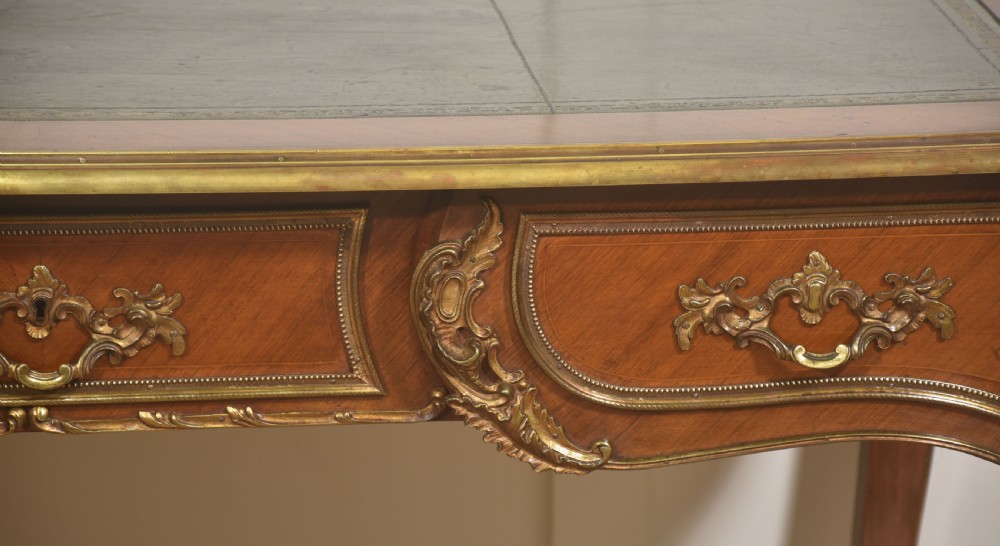 A French Louis XV style wooden bureau plat featuring three frieze drawers, gilt-bronze ormolu mounts, cabriole legs, and a green leather writing surface. View IV