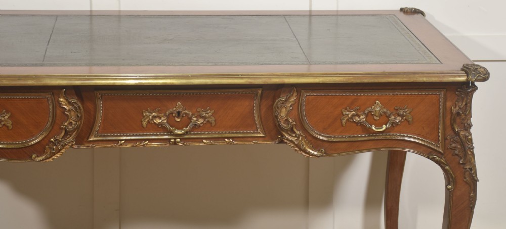 A French Louis XV style wooden bureau plat featuring three frieze drawers, gilt-bronze ormolu mounts, cabriole legs, and a green leather writing surface. View VI