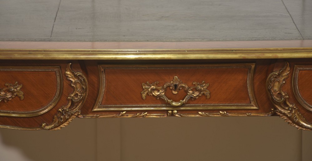 A French Louis XV style wooden bureau plat featuring three frieze drawers, gilt-bronze ormolu mounts, cabriole legs, and a green leather writing surface. View VII