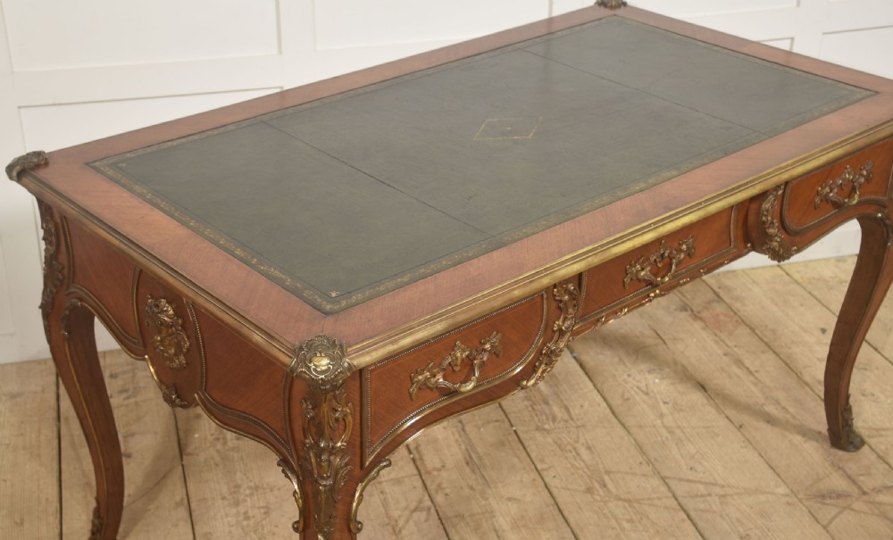 A French Louis XV style wooden bureau plat featuring three frieze drawers, gilt-bronze ormolu mounts, cabriole legs, and a green leather writing surface. View IX