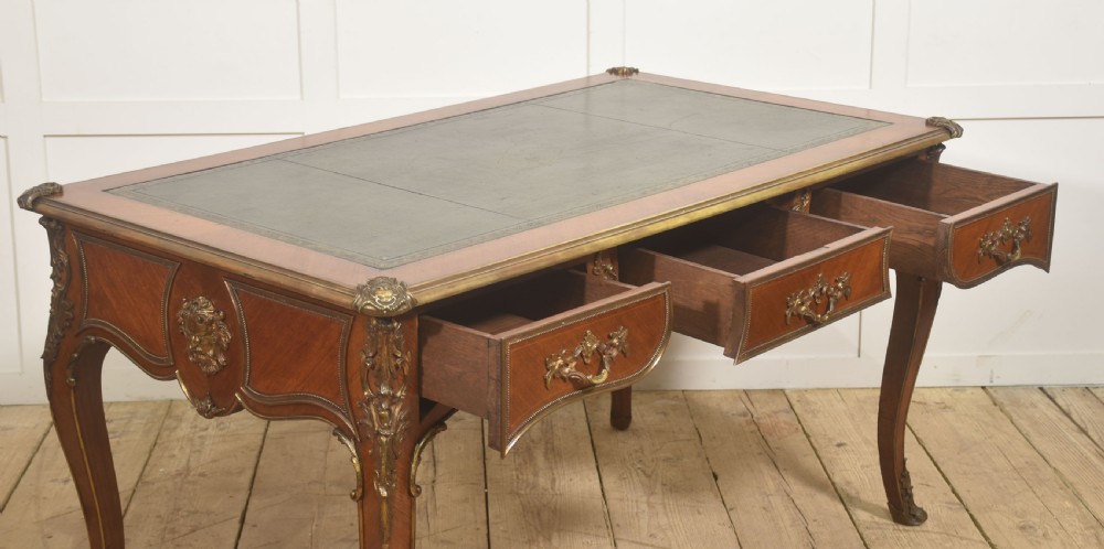 A French Louis XV style wooden bureau plat featuring three frieze drawers, gilt-bronze ormolu mounts, cabriole legs, and a green leather writing surface. View XV
