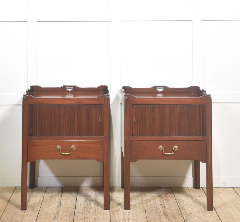 A pair of dark warm mahogany bedside cabinets featuring gallery tops with cutout handles, reeded tambour doors, and single lower drawers with brass handles.