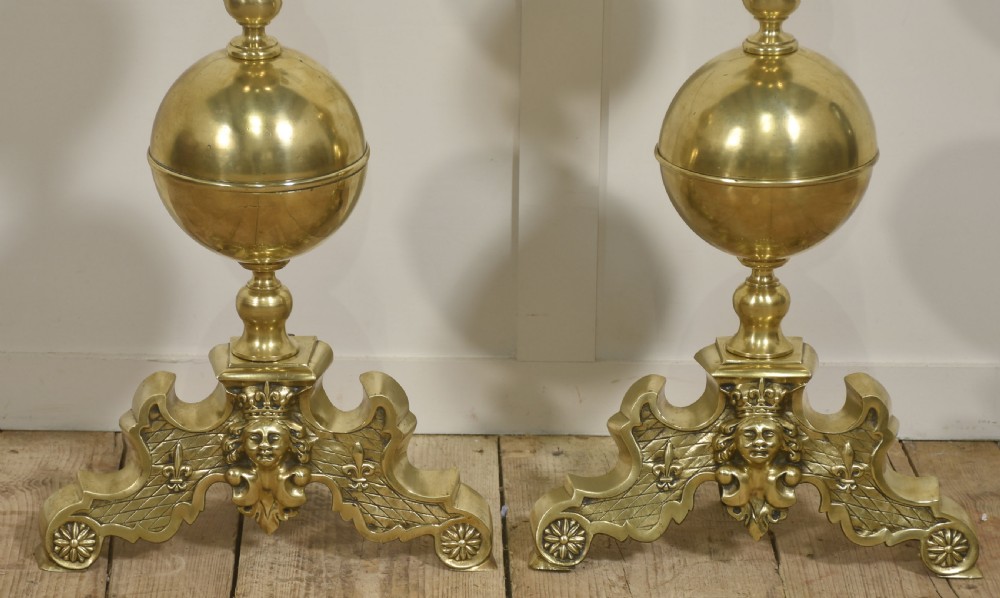 A pair of polished gilt brass andirons featuring double ball finials atop carved bases decorated with cherub faces, lattice work, and fleur-de-lis motifs. View VII