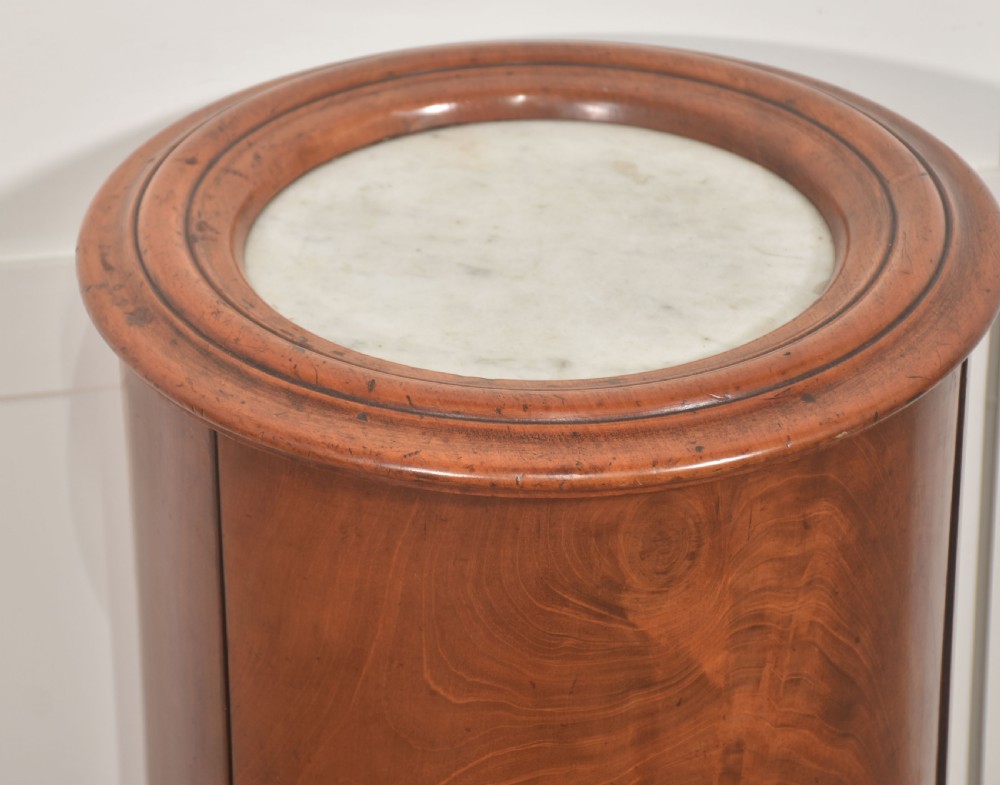 An Early Victorian cylindrical flame mahogany bedside cabinet featuring an inset marble top, a single curved door with a white ceramic knob, and a solid circular plinth base. View III