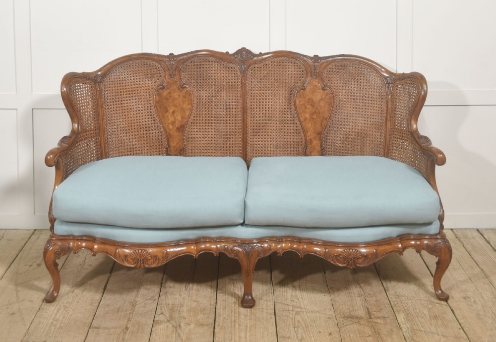 A Louis XV style carved walnut settee featuring a four-panel caned backrest, central burr walnut splats, light blue seat cushions, and carved cabriole legs.