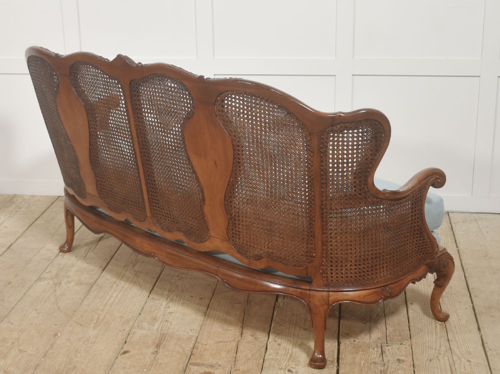 A Louis XV style carved walnut settee featuring a four-panel caned backrest, central burr walnut splats, light blue seat cushions, and carved cabriole legs. View X