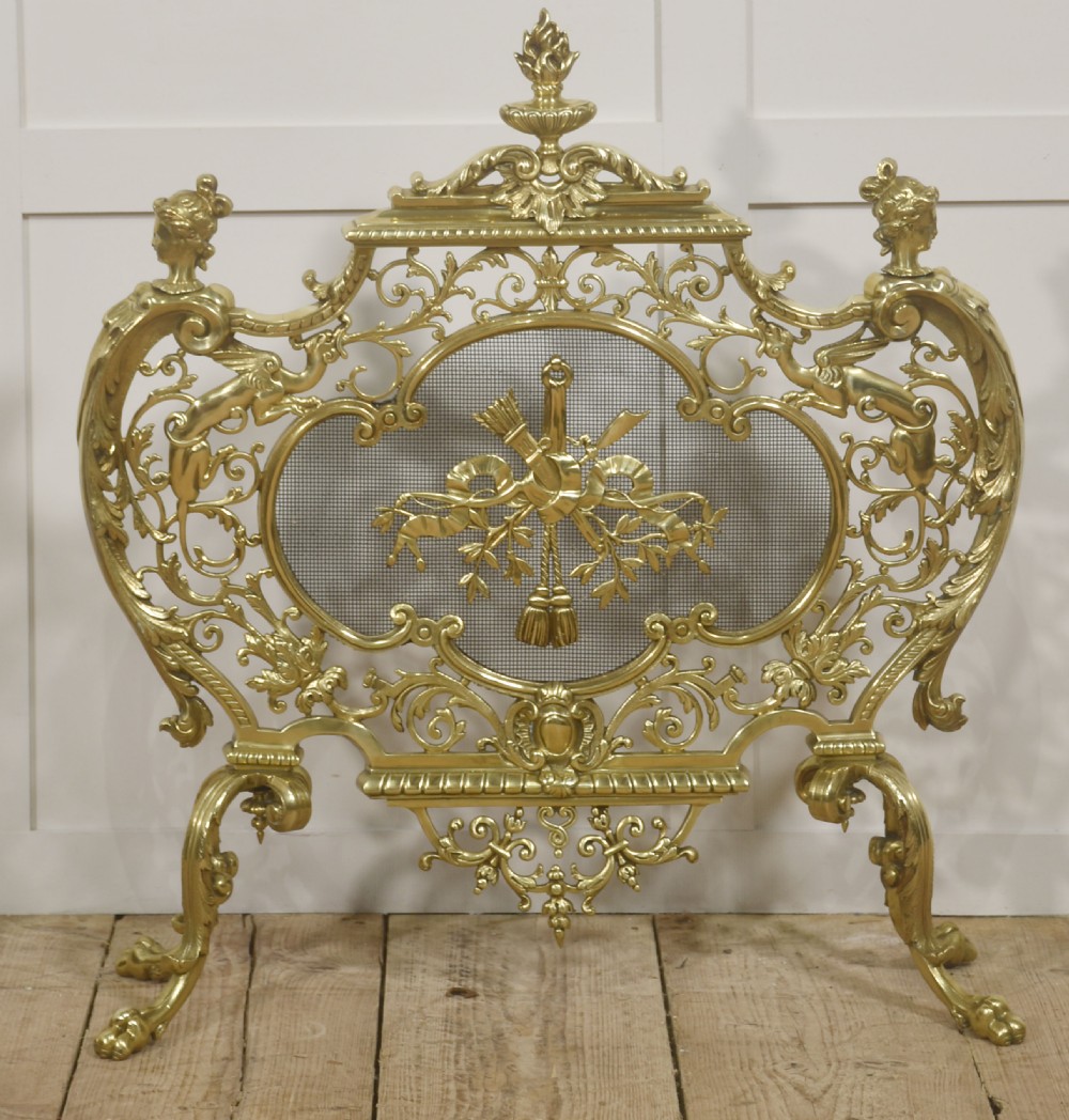 An ornate cast brass free-standing fire screen featuring pierced Rococo scrollwork, a mesh panel with torch and ribbon detailing, and scrolled feet.
