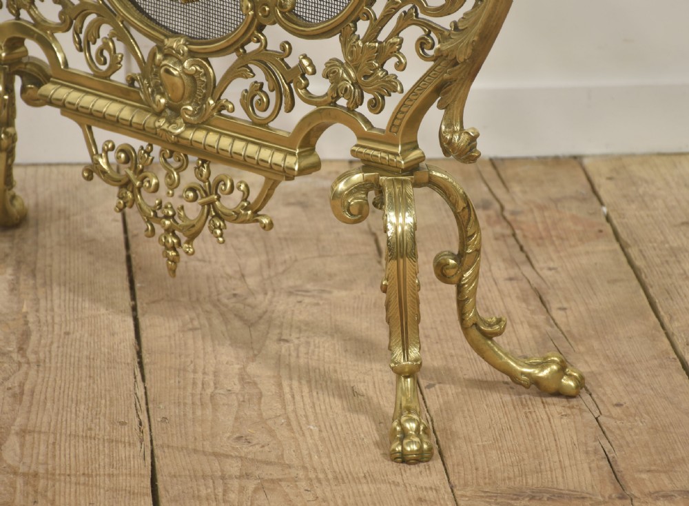 An ornate cast brass free-standing fire screen featuring pierced Rococo scrollwork, a mesh panel with torch and ribbon detailing, and scrolled feet. View IX
