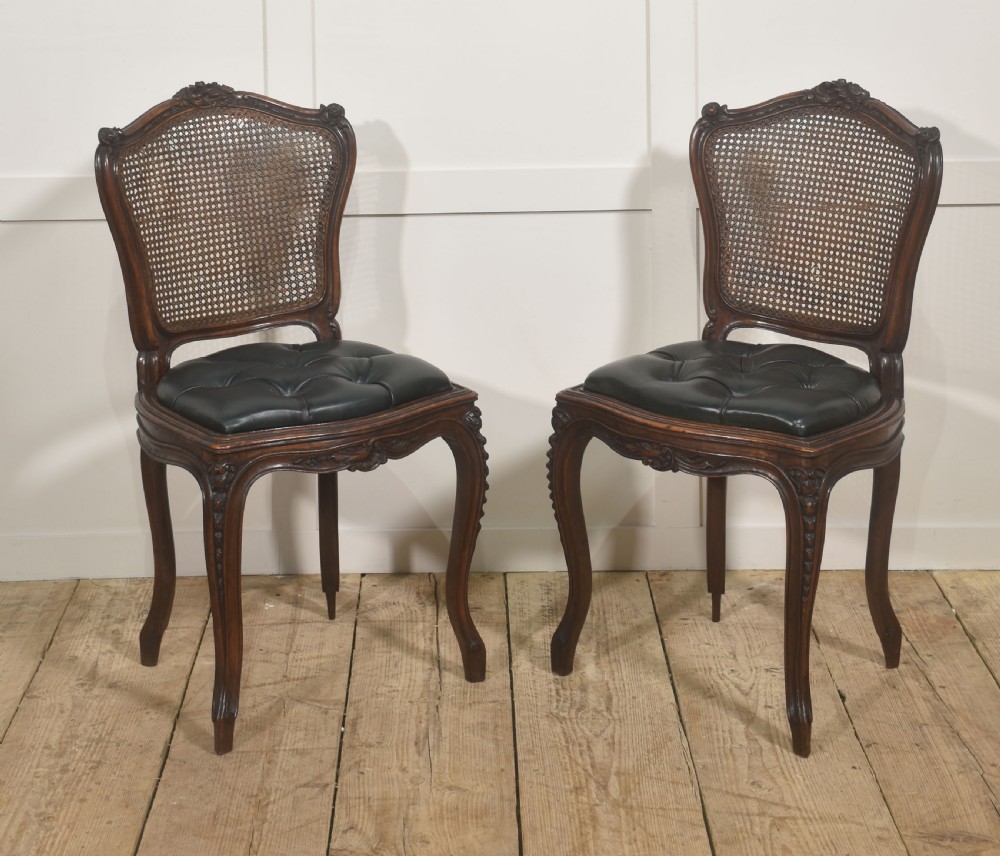 A pair of carved walnut side chairs in the Louis XV style, featuring woven cane backs, dark leather button-tufted seats, and cabriole legs.