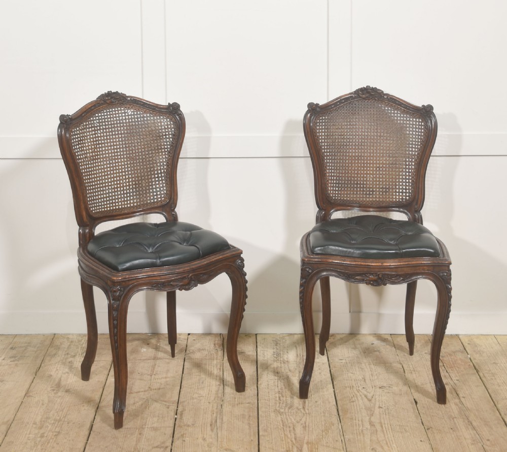 A pair of carved walnut side chairs in the Louis XV style, featuring woven cane backs, dark leather button-tufted seats, and cabriole legs. View V