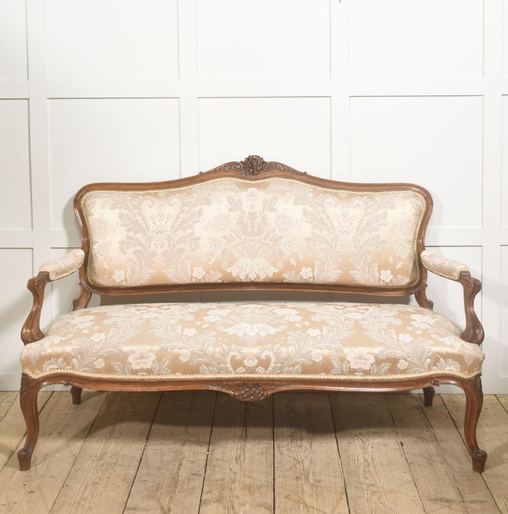 A Louis XV style carved walnut settee upholstered in beige damask fabric, featuring cabriole legs, padded armrests, and a carved cresting rail.