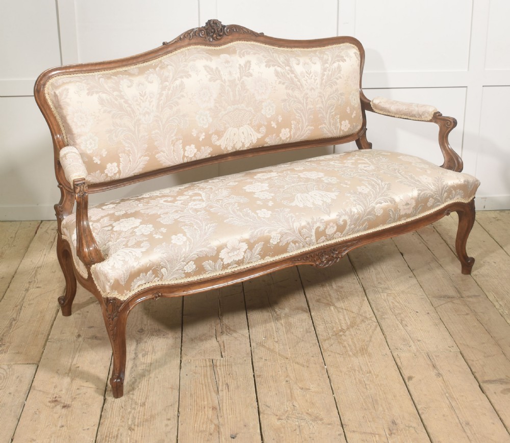 A Louis XV style carved walnut settee upholstered in beige damask fabric, featuring cabriole legs, padded armrests, and a carved cresting rail. View V