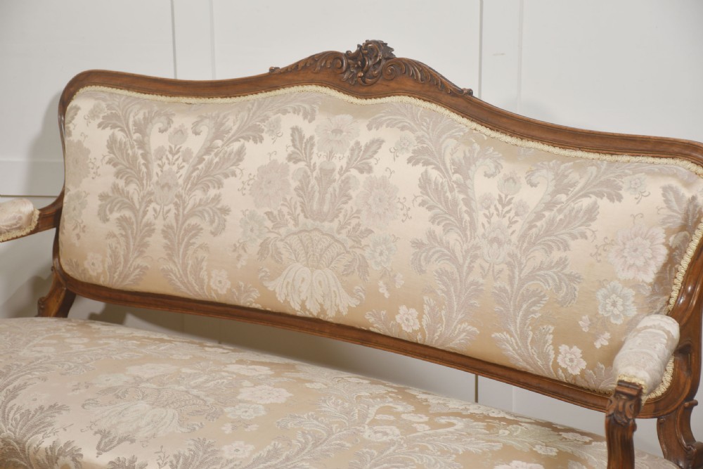 A Louis XV style carved walnut settee upholstered in beige damask fabric, featuring cabriole legs, padded armrests, and a carved cresting rail. View X