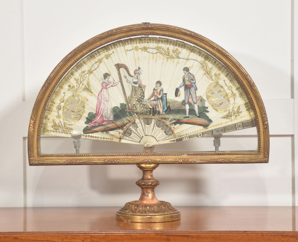 A hand-painted folding fan featuring a pastoral musical scene mounted inside a glazed giltwood frame on a turned pedestal.