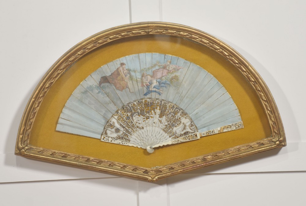 A nineteenth-century hand-painted fan depicting classical figures, mounted inside a fan-shaped gilt wood frame against an ochre background.