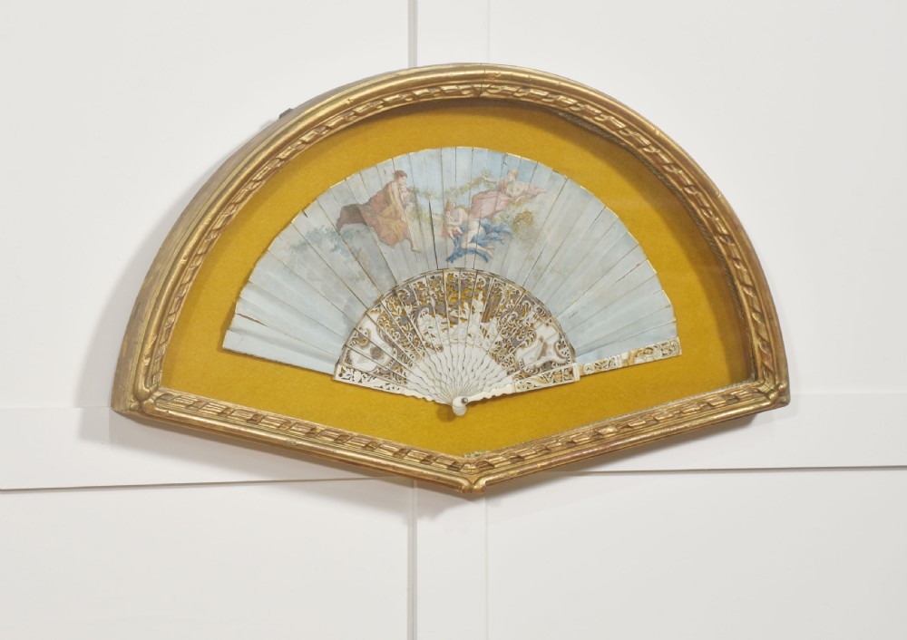 A nineteenth-century hand-painted fan depicting classical figures, mounted inside a fan-shaped gilt wood frame against an ochre background. View IV