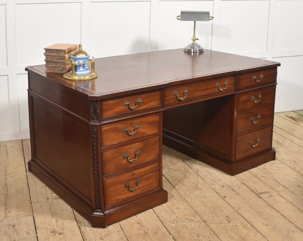 A large mahogany pedestal desk featuring a leather writing surface, multiple drawers with brass swan-neck handles, and carved corner details. View IV