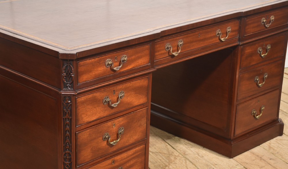 A large mahogany pedestal desk featuring a leather writing surface, multiple drawers with brass swan-neck handles, and carved corner details. View V