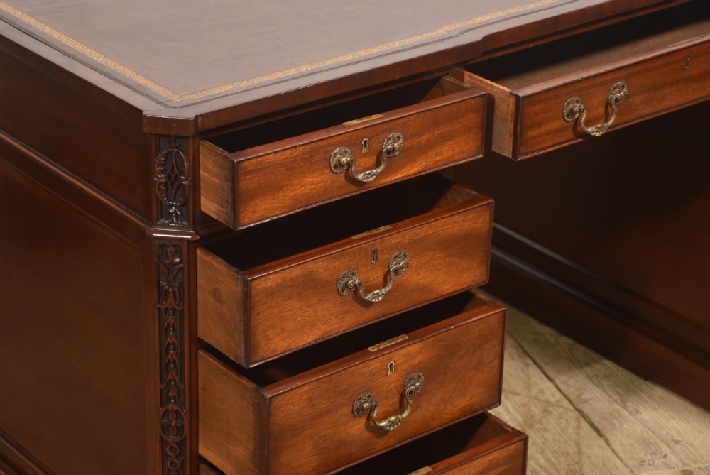 A large mahogany pedestal desk featuring a leather writing surface, multiple drawers with brass swan-neck handles, and carved corner details. View VII