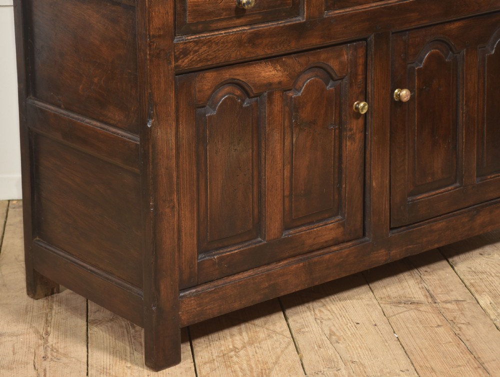 An antique dark oak Welsh dresser featuring an upper open shelf rack with two small side cupboards over a three-drawer base with two arched panel doors. View VI