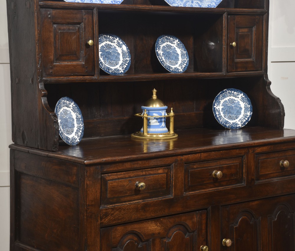 An antique dark oak Welsh dresser featuring an upper open shelf rack with two small side cupboards over a three-drawer base with two arched panel doors. View VII