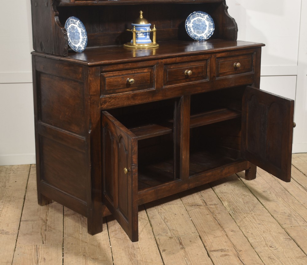 An antique dark oak Welsh dresser featuring an upper open shelf rack with two small side cupboards over a three-drawer base with two arched panel doors. View XI