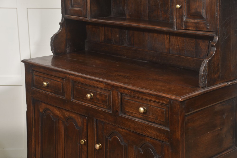 An antique dark oak Welsh dresser featuring an upper open shelf rack with two small side cupboards over a three-drawer base with two arched panel doors. View XVIII