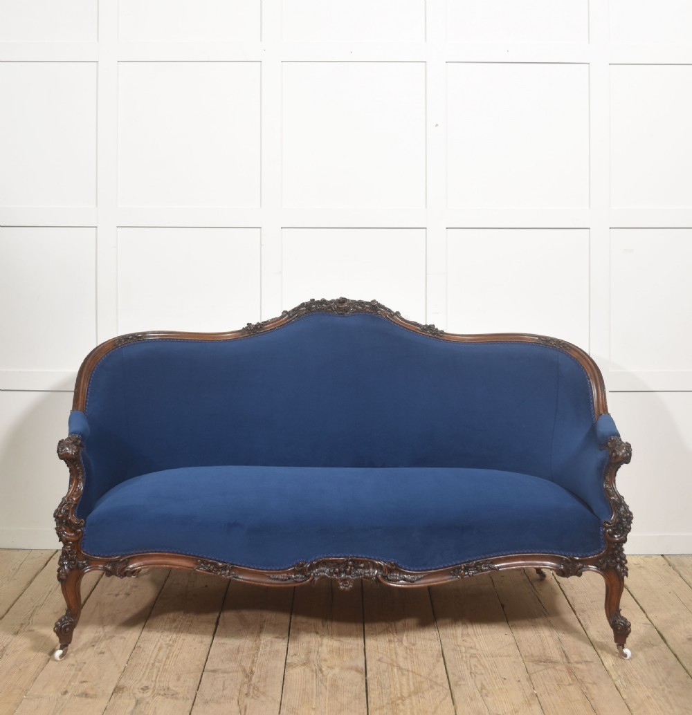 A 19th-century carved walnut settee featuring deep blue velvet upholstery, a serpentine crest rail, and hand-carved cabriole legs on small castors.
