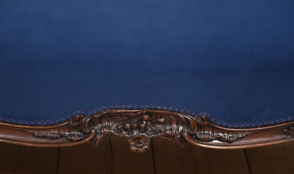 A 19th-century carved walnut settee featuring deep blue velvet upholstery, a serpentine crest rail, and hand-carved cabriole legs on small castors. View V