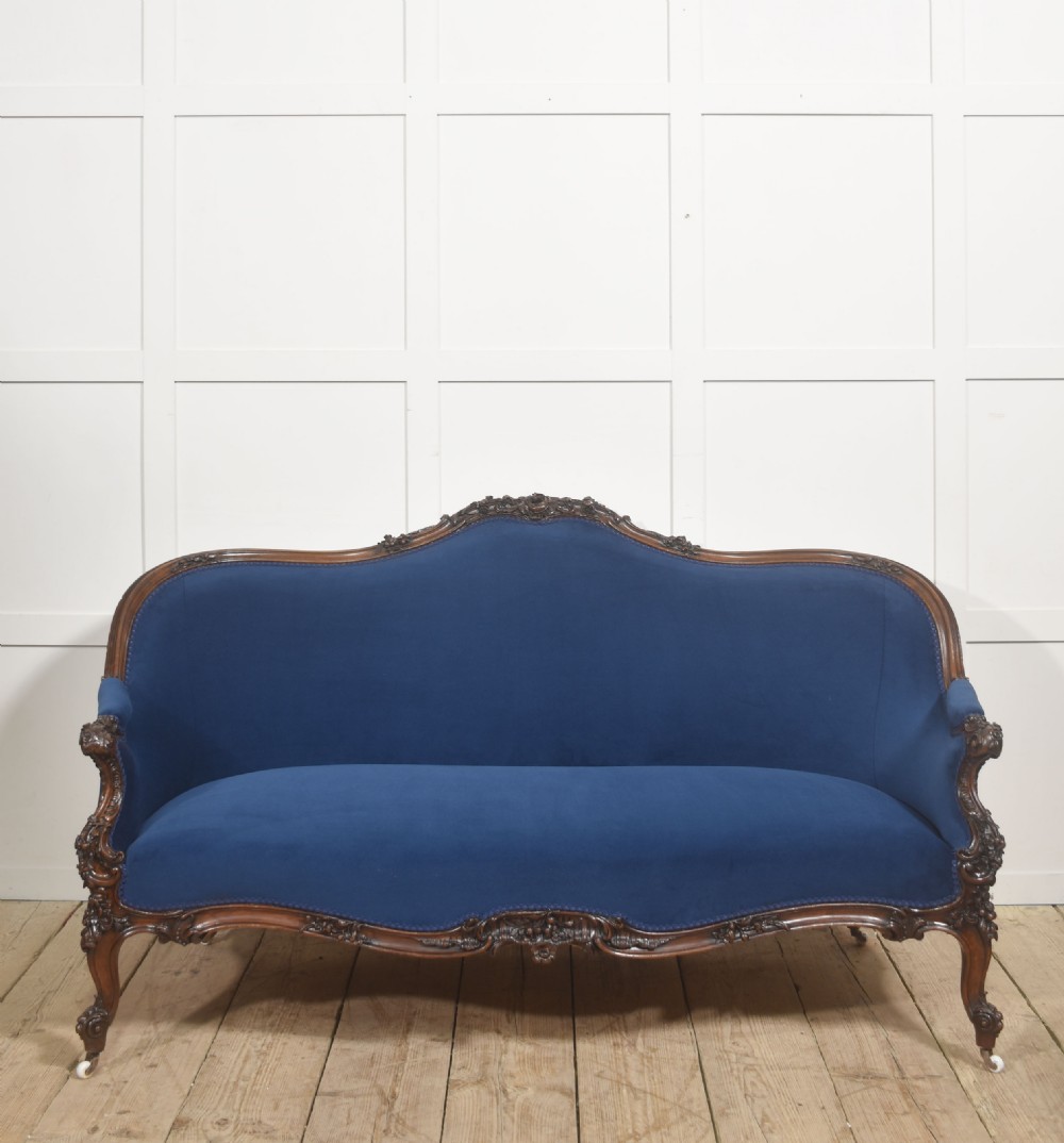 A 19th-century carved walnut settee featuring deep blue velvet upholstery, a serpentine crest rail, and hand-carved cabriole legs on small castors. View VI