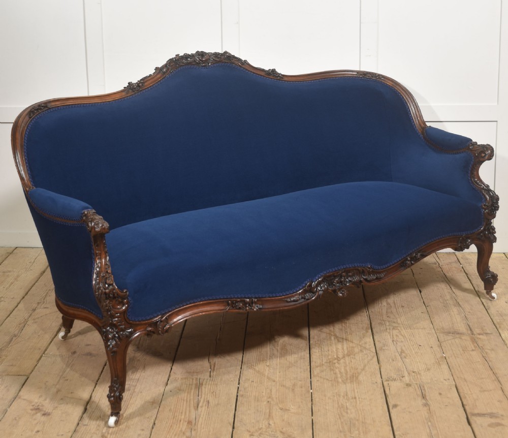 A 19th-century carved walnut settee featuring deep blue velvet upholstery, a serpentine crest rail, and hand-carved cabriole legs on small castors. View VIII