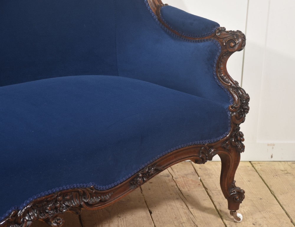 A 19th-century carved walnut settee featuring deep blue velvet upholstery, a serpentine crest rail, and hand-carved cabriole legs on small castors. View X