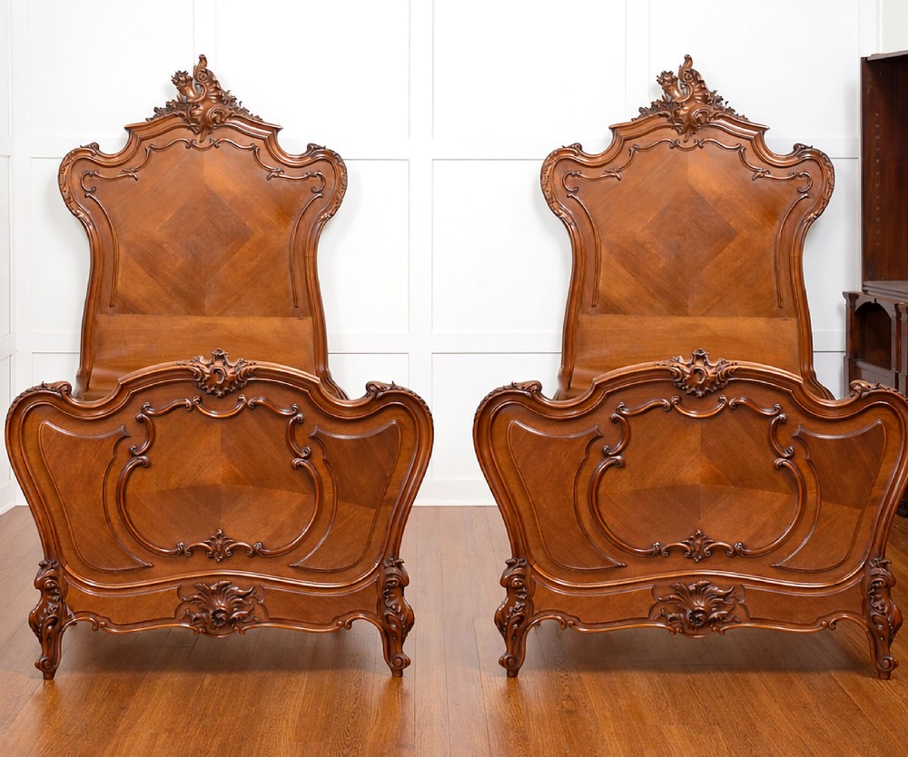 A matching pair of carved walnut single bed frames with high crest headboards, serpentine footboards, and book-matched timber panels.