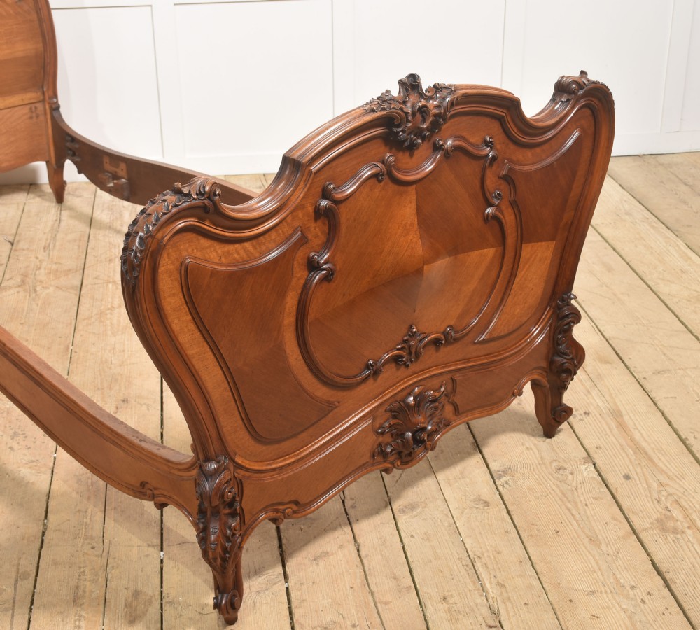 A matching pair of carved walnut single bed frames with high crest headboards, serpentine footboards, and book-matched timber panels. View IX