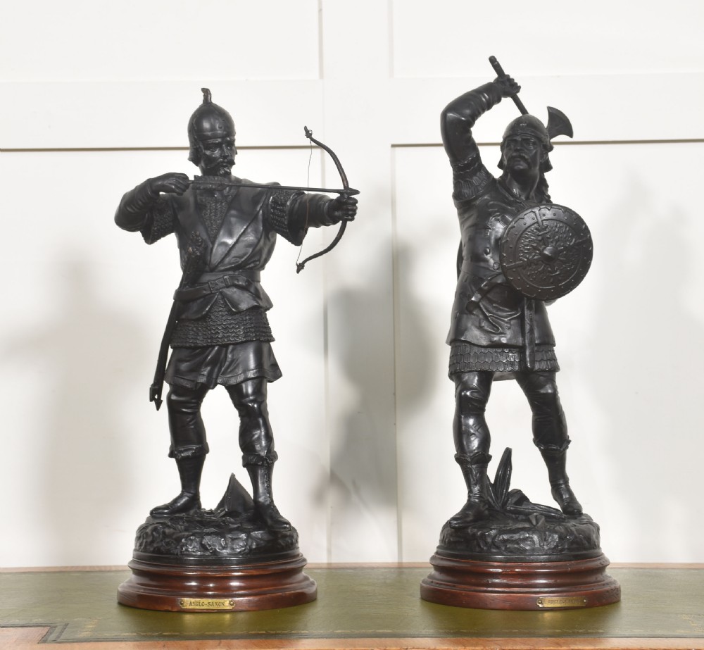 A pair of dark bronzed metal figures depicting Anglo-Saxon warriors, one drawn with a bow and the other raising an axe, mounted on circular wooden bases.