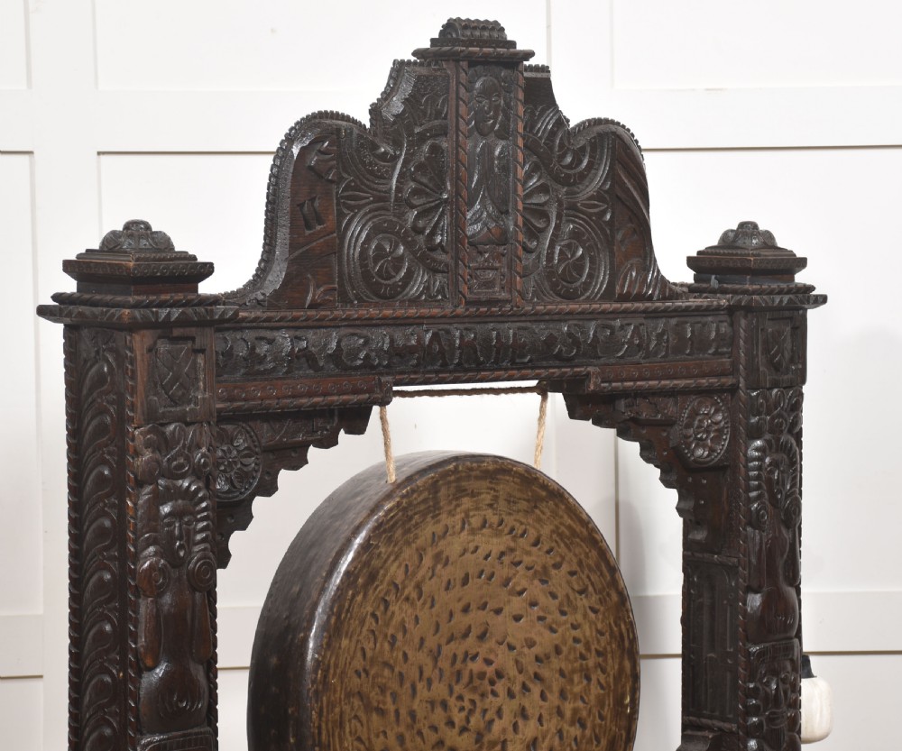 A large dark oak freestanding frame decorated with carved human masks and foliate relief, holding a central circular hammered brass gong with an accompanying padded striker. View II