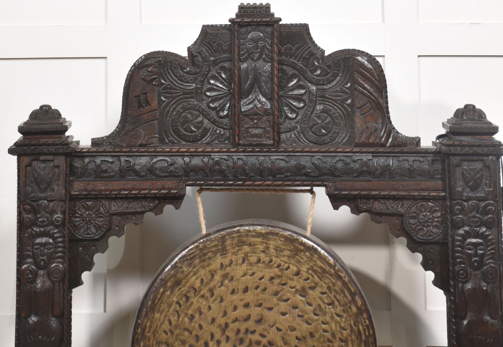 A large dark oak freestanding frame decorated with carved human masks and foliate relief, holding a central circular hammered brass gong with an accompanying padded striker. View V