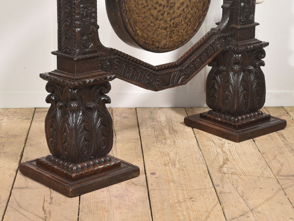 A large dark oak freestanding frame decorated with carved human masks and foliate relief, holding a central circular hammered brass gong with an accompanying padded striker. View VII