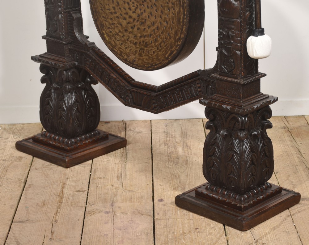 A large dark oak freestanding frame decorated with carved human masks and foliate relief, holding a central circular hammered brass gong with an accompanying padded striker. View X