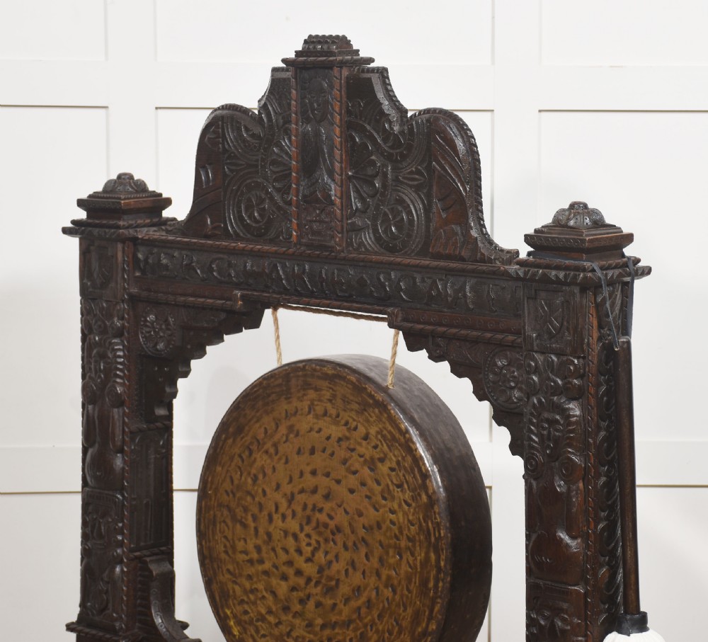 A large dark oak freestanding frame decorated with carved human masks and foliate relief, holding a central circular hammered brass gong with an accompanying padded striker. View XI