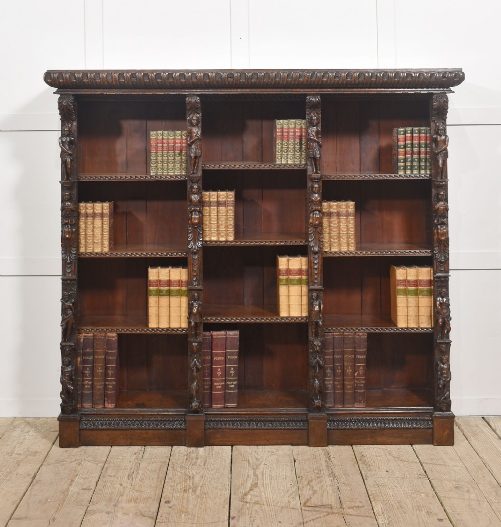 A late 19th-century carved oak open bookcase featuring three vertical bays, carved figures along the stiles, and decorative egg-and-dart moulding along the top cornice.