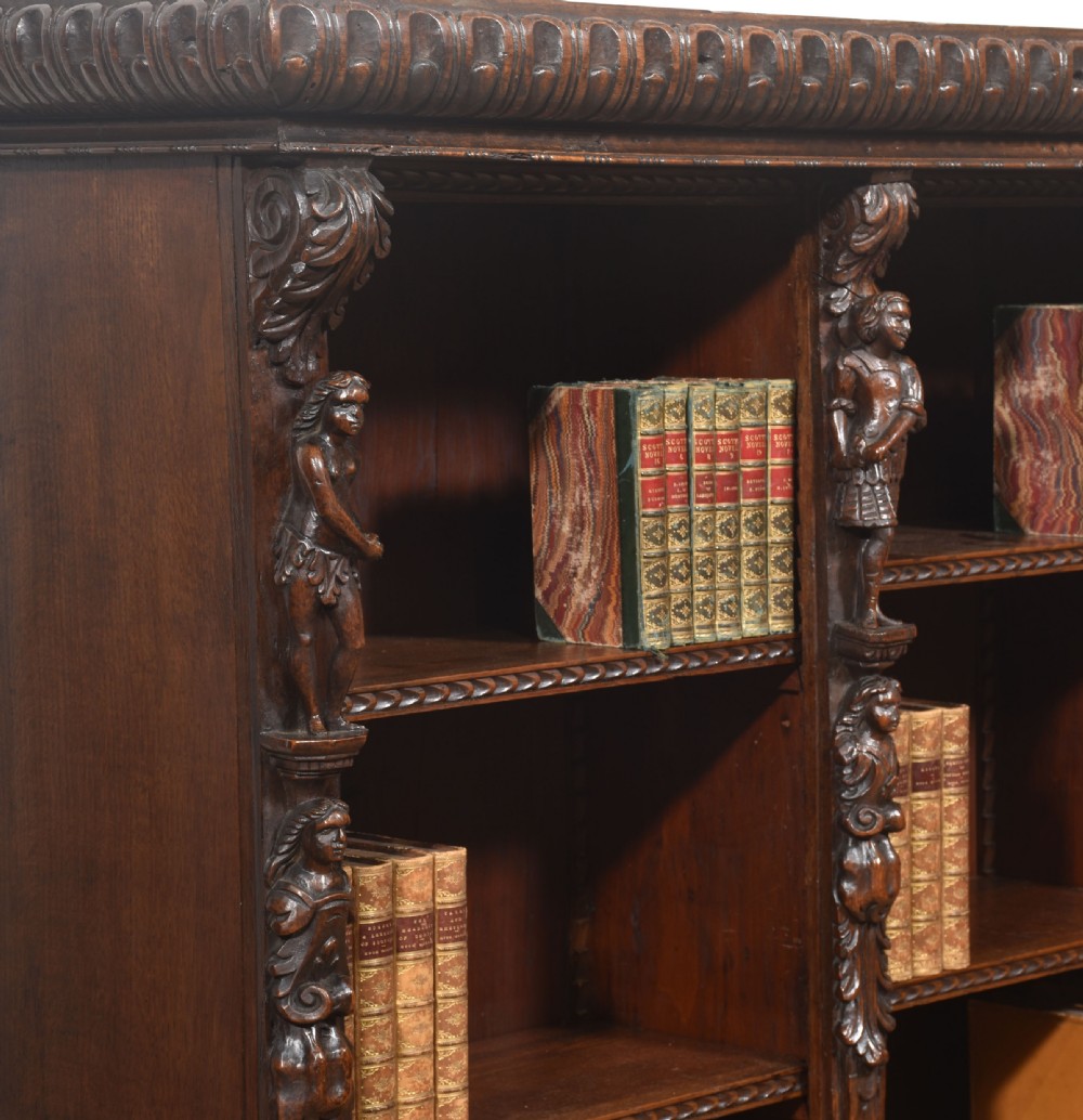 A late 19th-century carved oak open bookcase featuring three vertical bays, carved figures along the stiles, and decorative egg-and-dart moulding along the top cornice. View VIII