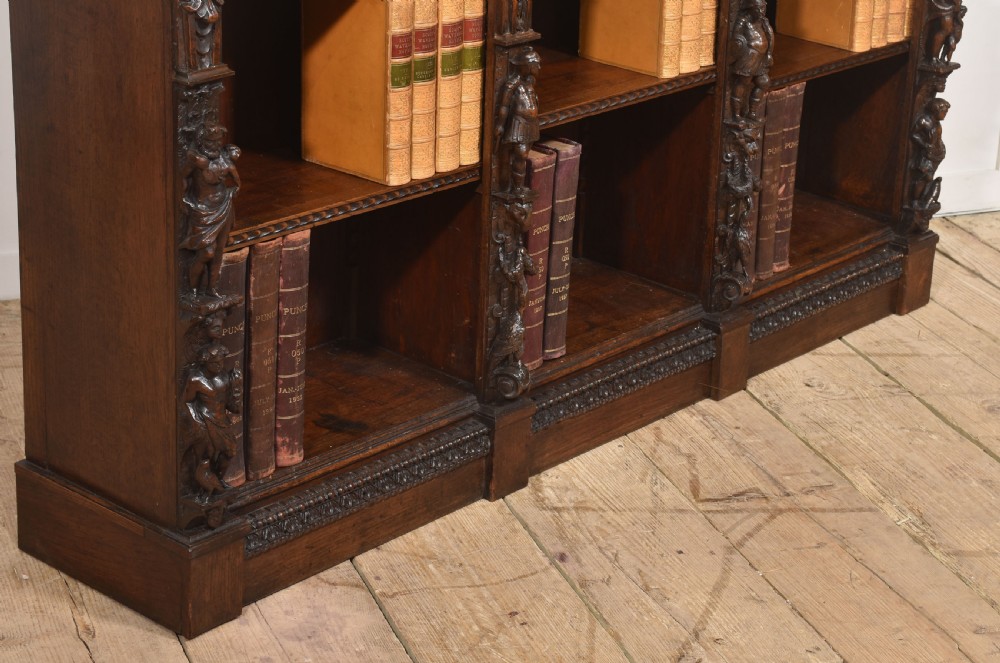 A late 19th-century carved oak open bookcase featuring three vertical bays, carved figures along the stiles, and decorative egg-and-dart moulding along the top cornice. View X