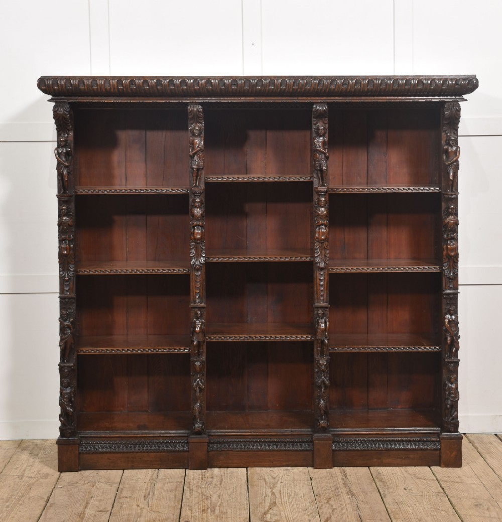 A late 19th-century carved oak open bookcase featuring three vertical bays, carved figures along the stiles, and decorative egg-and-dart moulding along the top cornice. View XI