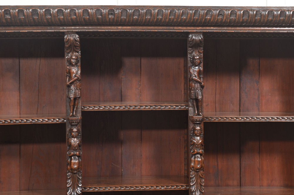 A late 19th-century carved oak open bookcase featuring three vertical bays, carved figures along the stiles, and decorative egg-and-dart moulding along the top cornice. View XII
