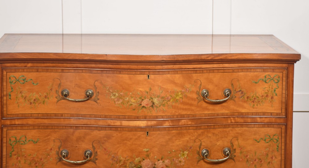 A Sheraton Revival satinwood chest of drawers featuring a serpentine front, three drawers decorated with painted floral swags, and brass handles. View II